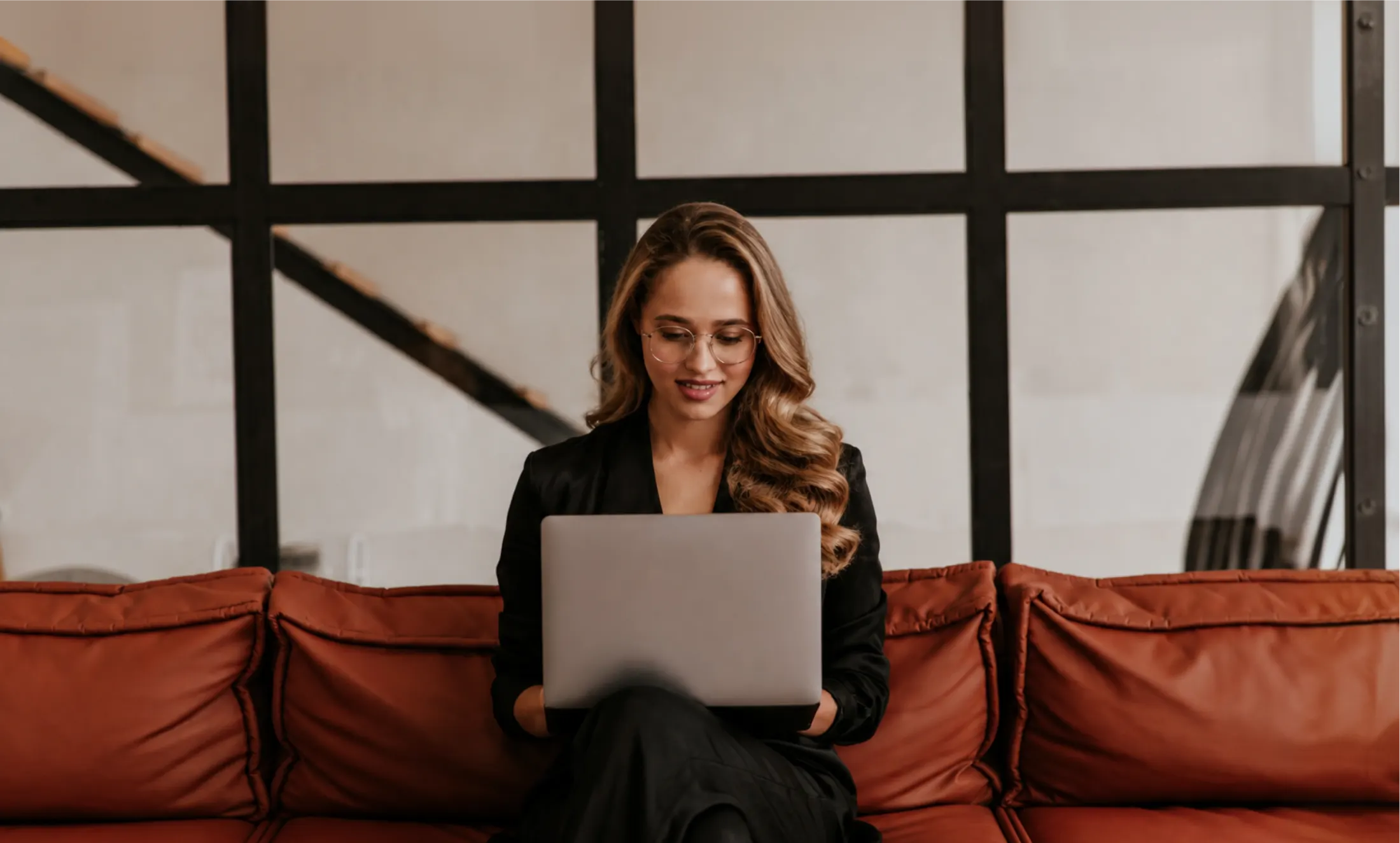 A woman in glasses smiling at her laptop on a leather sofa